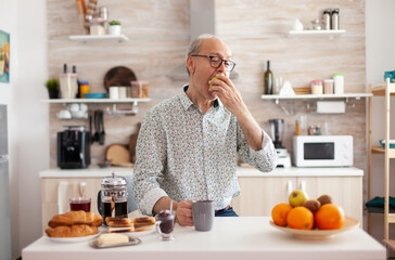 Fototapeta na wymiar Elderly retired person biting apple during breakfast in mdoern cozy kitchen holding cup of coffee. Authentic portrait of senior man enjoying for healthy green happy fresh lifestyle with organic food.