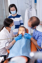Little girl getting treatment for caries from dentist and assistant wearing dental bib. Mother with her kid in stomatology clinic for teeth examine using modern instruments.