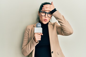 Young hispanic woman holding reporter microphone stressed and frustrated with hand on head,...