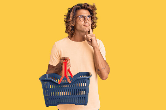 Young Hispanic Man Holding Supermarket Shopping Basket Serious Face Thinking About Question With Hand On Chin, Thoughtful About Confusing Idea