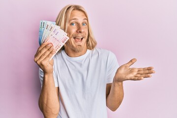 Caucasian young man with long hair holding new taiwan dollars banknotes celebrating achievement with happy smile and winner expression with raised hand