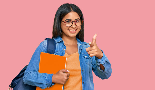 Young latin girl wearing student backpack and holding books pointing fingers to camera with happy and funny face. good energy and vibes.