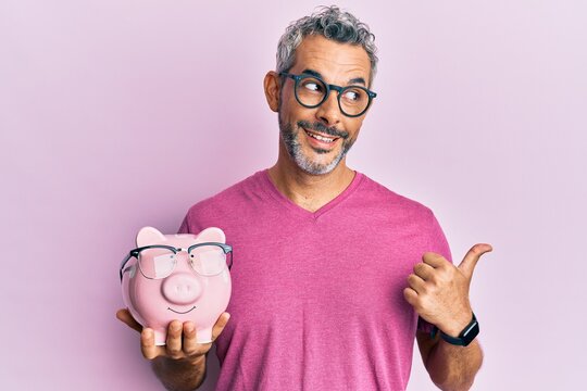 Middle age grey-haired man holding piggy bank with glasses pointing thumb up to the side smiling happy with open mouth
