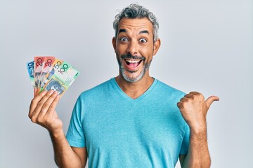 Middle age grey-haired man holding australian dollars pointing thumb up to the side smiling happy with open mouth