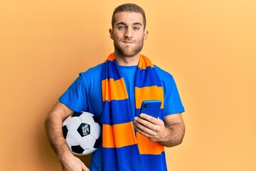 Young caucasian man holding football ball looking at smartphone puffing cheeks with funny face. mouth inflated with air, catching air.