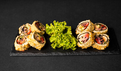 Custom sushi roll in tempura with nori, fresh salmon, tuna, avocado, masago caviar, drizzled with pineapple sauce with salad pouring as decoration on a black plate on a black table and background.