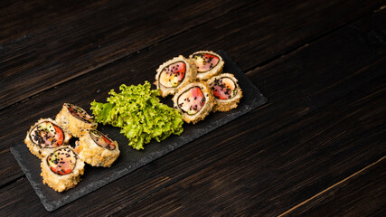 Custom sushi roll in tempura with nori, fresh salmon, tuna, avocado, masago caviar, drizzled with pineapple sauce with salad pouring as decoration on a black plate on a wooden table and background.