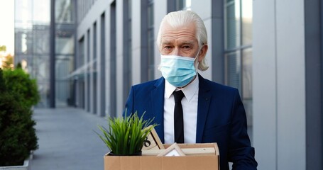 Portrait of old retired Caucasian gray-haired man in mask standing outdoor and holding box with...