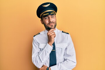 Handsome hispanic man wearing airplane pilot uniform looking confident at the camera smiling with crossed arms and hand raised on chin. thinking positive. © Krakenimages.com