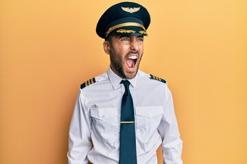 Handsome hispanic man wearing airplane pilot uniform angry and mad screaming frustrated and furious, shouting with anger. rage and aggressive concept. - obrazy, fototapety, plakaty