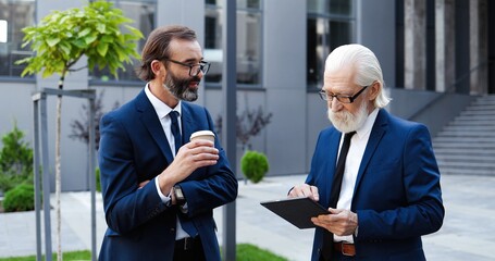 Two Caucasian senior businessmen standing outdoor in city center, drinking coffee and working on tablet device. Men in suits and ties colleagues discussing business and job with gadget computer.