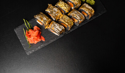 Custom sushi roll with nori, cuttlefish ink, fresh salmon, cream cheese, pepper pumpkin, eel, unagi sauce and edible gold leaf with red ginger, wasabi on a square plate on black table and background.