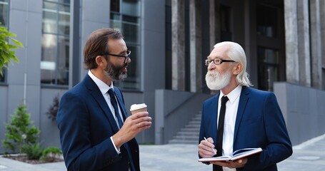 Two Caucasian senior businessmen standing outdoor in city center, drinking coffee and working in notebook. Men in suits and ties colleagues discussing business and job with planner and pen in hands.