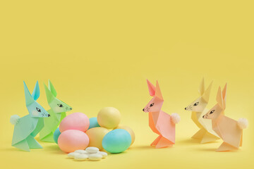 Paper Easter bunnies-origami made of colored paper and colored eggs on a delicate yellow background. The concept of the celebration of Easter, greeting card, crafts with your own hands.