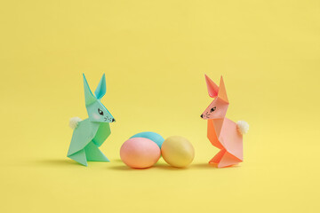 Paper Easter bunnies-origami made of colored paper and colored eggs on a delicate yellow background. The concept of the celebration of Easter, greeting card, crafts with your own hands.