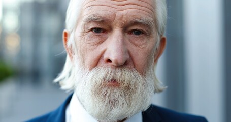 Portrait of Caucasian old gray-haired man standing outside and looking at camera with sad eyes....
