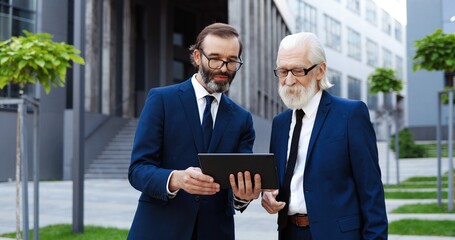 Two Caucasian senior elegant businessmen in glasses and ties standing outside at street and...