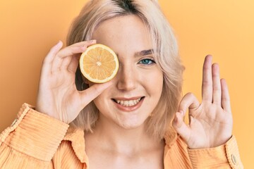 Young blonde girl holding lemon over eye doing ok sign with fingers, smiling friendly gesturing excellent symbol
