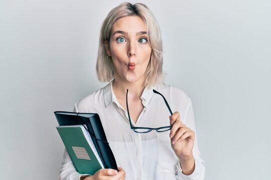 Young blonde girl reading a book and holding glasses making fish face with mouth and squinting eyes, crazy and comical.