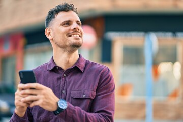 Young hispanic man smiling happy using smartphone at the city.