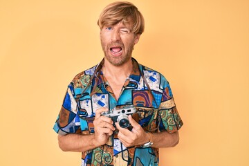 Young blond man holding vintage camera winking looking at the camera with sexy expression, cheerful...