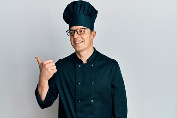 Handsome young man wearing professional cook uniform and hat pointing thumb up to the side smiling happy with open mouth