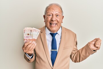 Senior caucasian man holding 10 colombian pesos banknotes screaming proud, celebrating victory and success very excited with raised arm