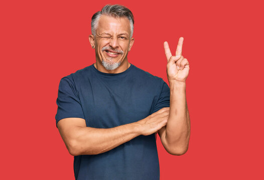 Middle age grey-haired man wearing casual clothes smiling with happy face winking at the camera doing victory sign. number two.
