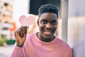 Young african american man smiling happy holding heart paper at the city.
