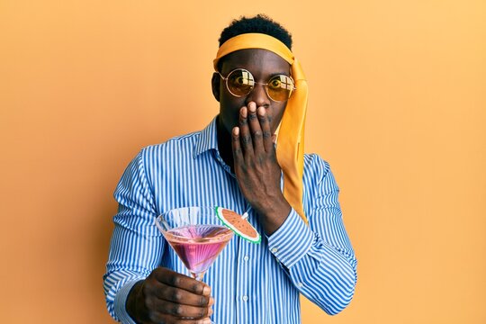 Handsome black man drunk wearing tie over head and sunglasses drinking a cocktail covering mouth with hand, shocked and afraid for mistake. surprised expression