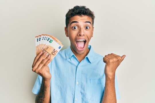 Young handsome african american man holding bunch of 50 euro banknotes pointing thumb up to the side smiling happy with open mouth