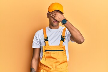 Young handsome african american man wearing handyman uniform over yellow background covering eyes with hand, looking serious and sad. sightless, hiding and rejection concept