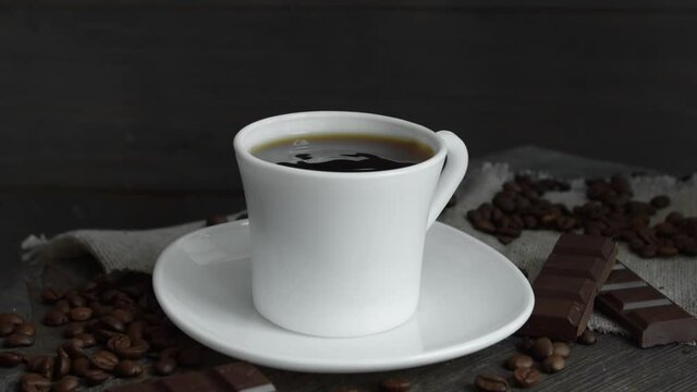 A drop of coffee falls into a white mug of espresso with coffee beans scattered on the table and chocolate on a wooden table. Fresh delicious coffee is ready to drink.