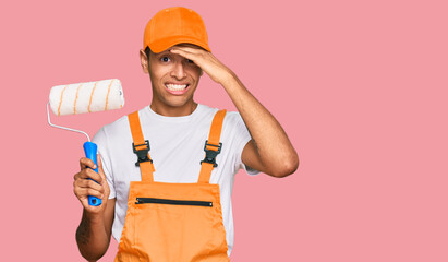 Young handsome african american man wearing cap and painter clothes holding painting roll stressed and frustrated with hand on head, surprised and angry face