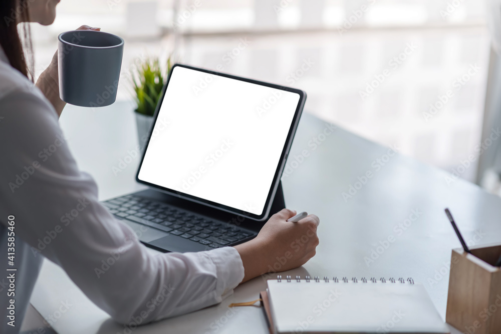 Wall mural Young woman drinking coffee and using tablet in the office. Blank white screen. Mock up.