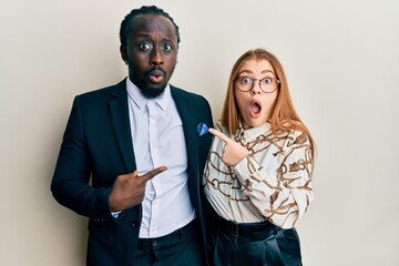 Young interracial couple wearing business and elegant clothes surprised pointing with finger to the...