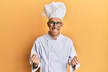 Bald man with beard wearing professional cook uniform very happy and excited doing winner gesture with arms raised, smiling and screaming for success. celebration concept.