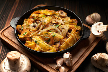 Homemade fried potatoes, with onions, mushrooms and garlic, in a frying pan, on a wooden board