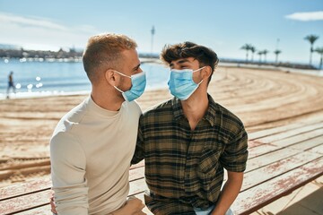 Young gay couple wearing medical mask sitting on the bench at the beach.