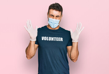 Hispanic young man wearing medical mask and volunteer t shirt celebrating mad and crazy for success with arms raised and closed eyes screaming excited. winner concept