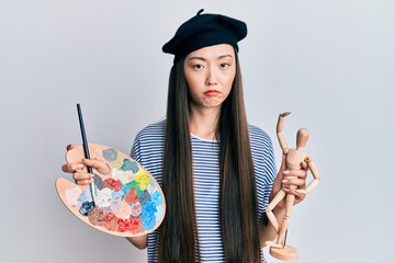 Young chinese woman wearing artist look with beret holding manikin depressed and worry for distress, crying angry and afraid. sad expression.