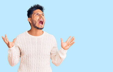 Fototapeta premium Handsome latin american young man wearing casual winter sweater crazy and mad shouting and yelling with aggressive expression and arms raised. frustration concept.