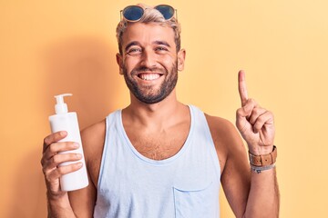 Young handsome blond man with beard on vacation holding bottle of sunscreen to protect skin smiling with an idea or question pointing finger with happy face, number one