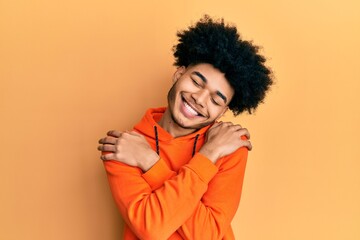 Young african american man with afro hair wearing casual sweatshirt hugging oneself happy and...