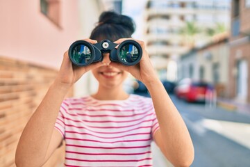 Young hispanic girl smiling happy looking for new opportunity using binoculars at the city.