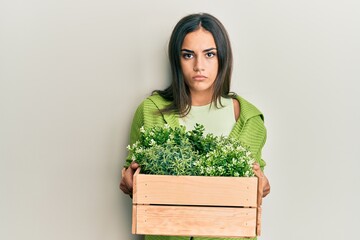 Young brunette woman holding wooden plant pot relaxed with serious expression on face. simple and natural looking at the camera.