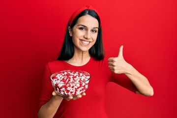 Young hispanic woman holding bowl of pills smiling happy and positive, thumb up doing excellent and approval sign