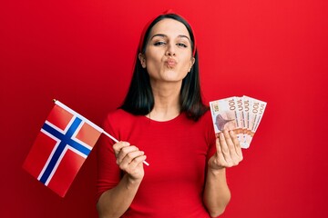 Young hispanic woman holding norway flag and krone banknotes looking at the camera blowing a kiss being lovely and sexy. love expression.