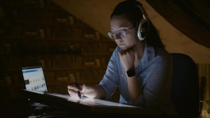 Portrait of female graphic designer working in dark room at computer. Young woman working for herself draws on graphic tablet, listens to music on headphones - Powered by Adobe