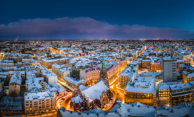 Obraz premium Riga city panorama in winter time. Street lights glowing in blue hour. Snowy rooftops. Iconic buildings and architecture. Drone footage. 